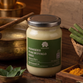 A jar of FARMSEED Pure Buffalo Ghee sits on a rustic wooden table surrounded by cooking ingredients and brass utensils. The jar has a green label and a silver lid, with warm, ambient lighting in the background.